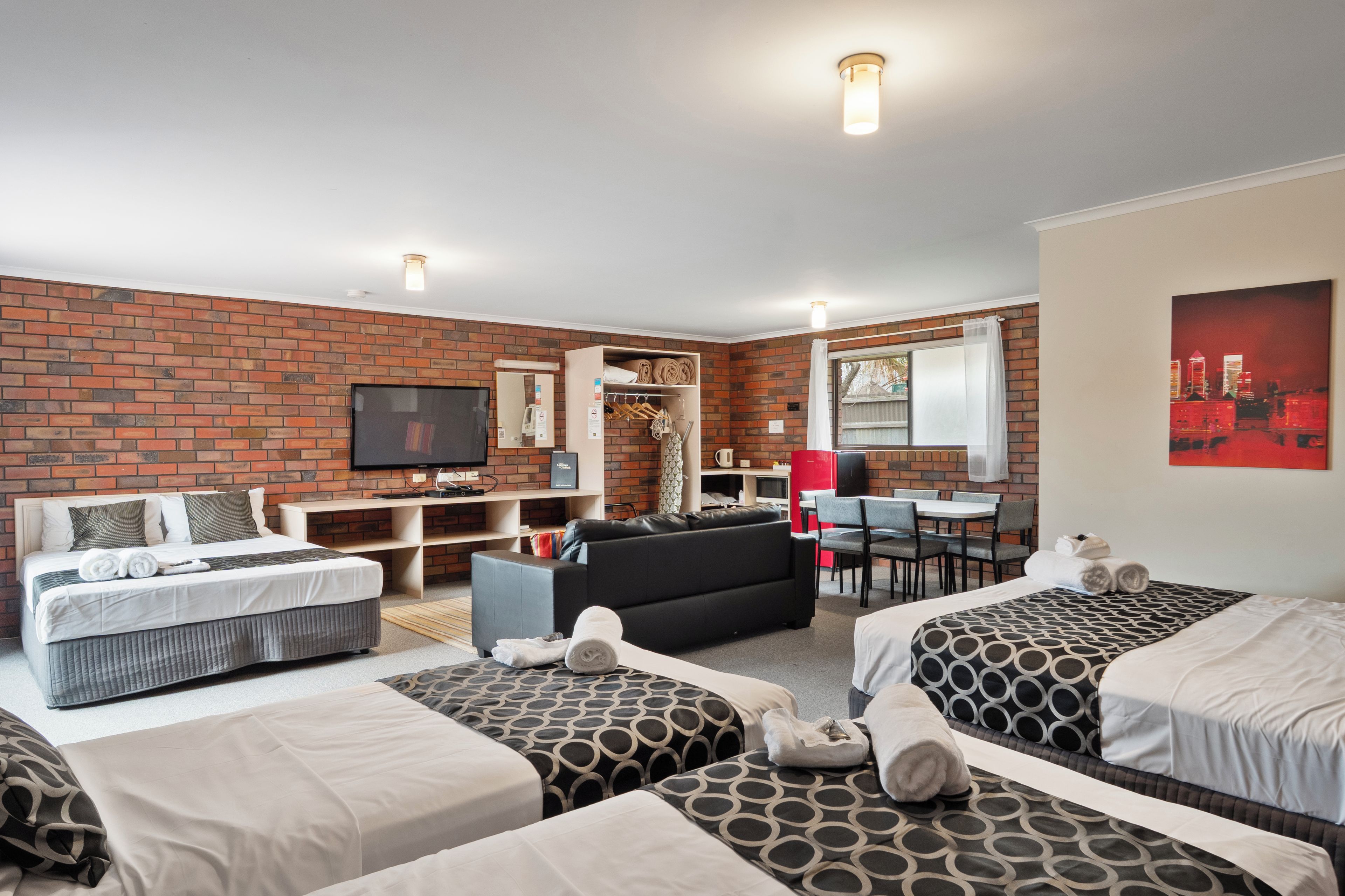 A hotel room with a brick wall, a flat-screen TV, a sofa, a dining table with chairs, and two beds with patterned bedding.