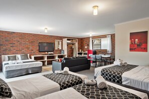 A hotel room with a brick wall, a flat-screen TV, a sofa, a dining table with chairs, and two beds with patterned bedding. - The Coorrabin Inn (Stawell)