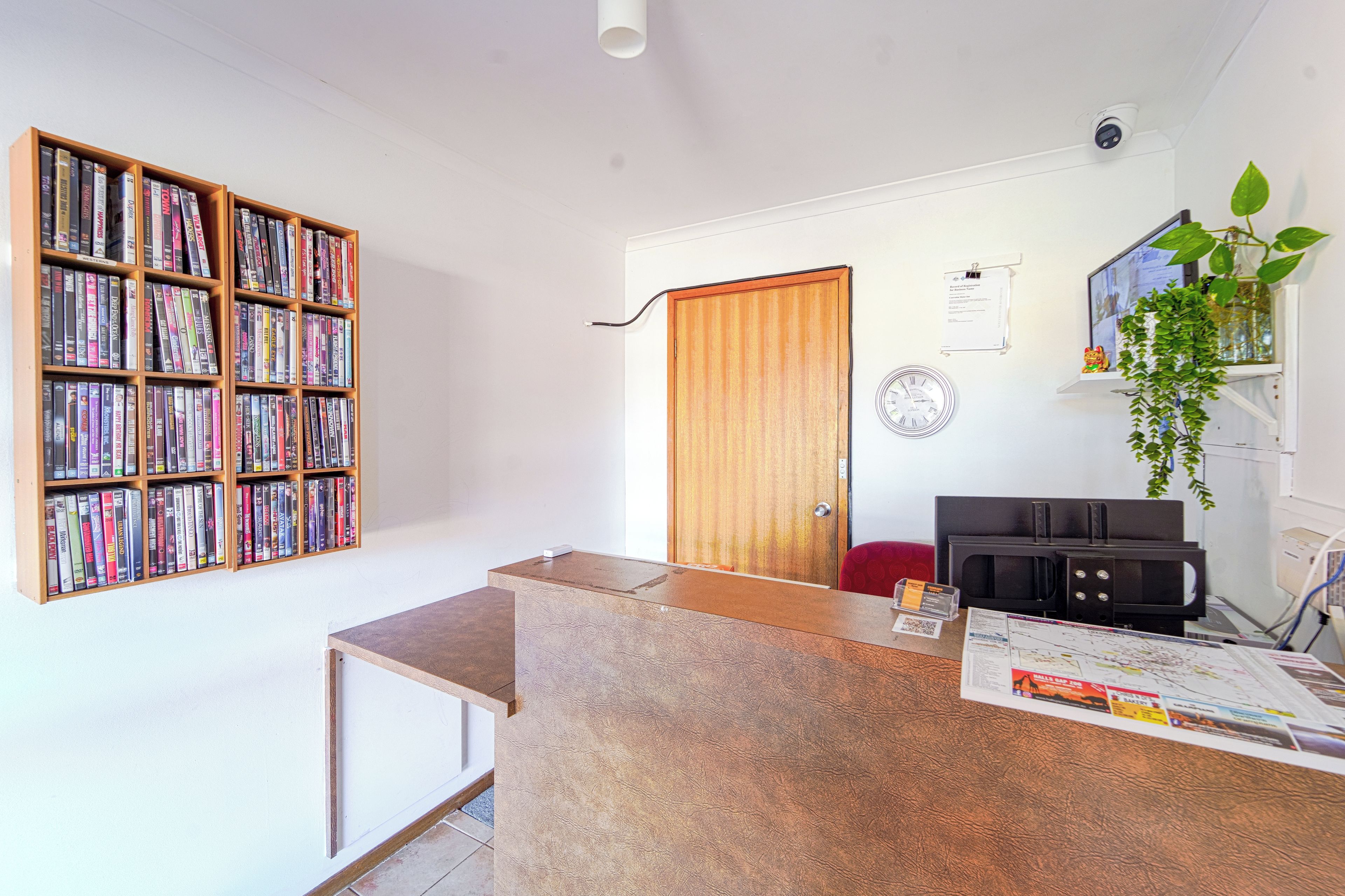 Una habitación con una estantería de madera llena de libros, un mostrador de recepción de madera, una puerta, un reloj en la pared y una planta en maceta.