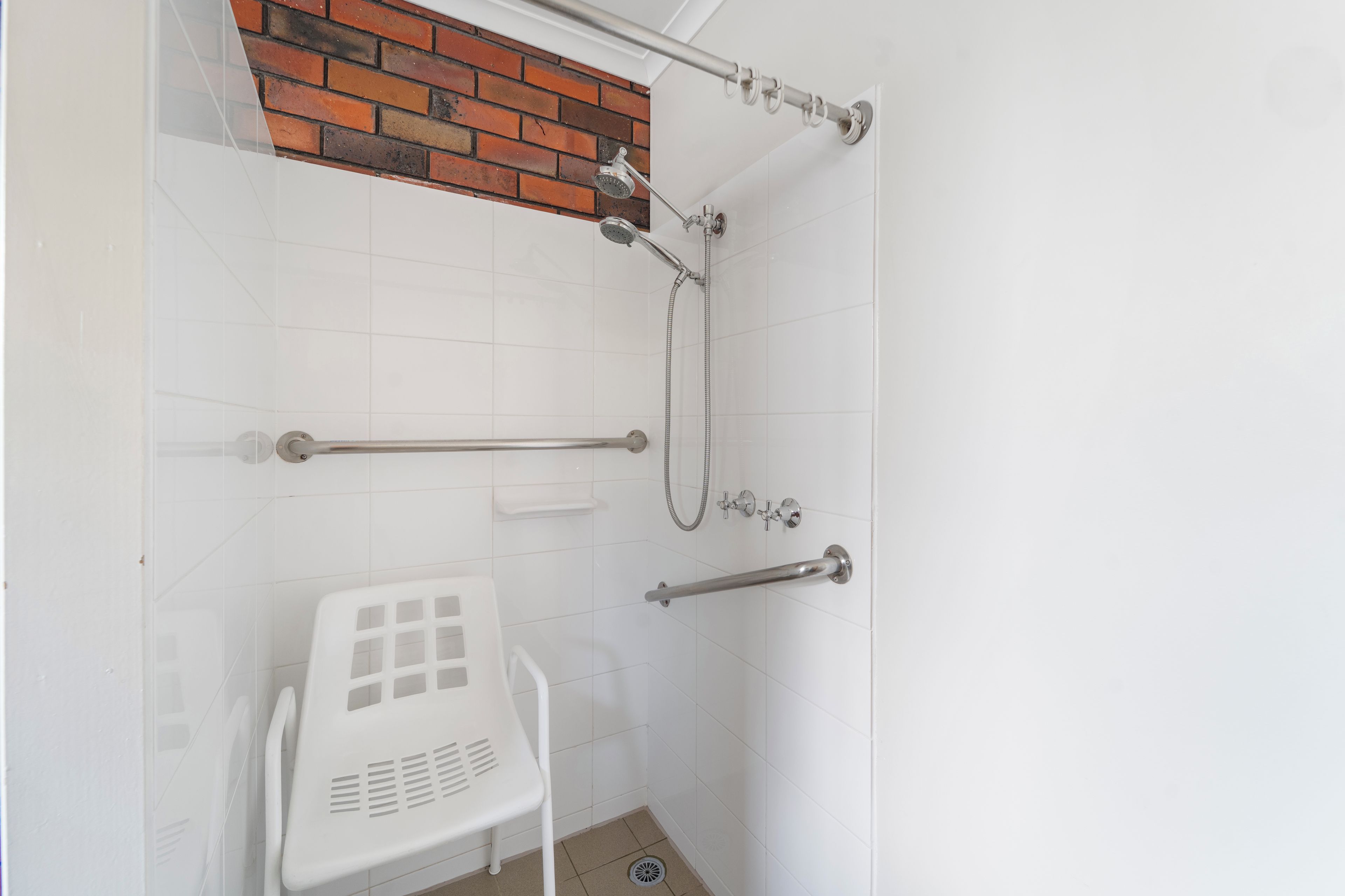 Un espace douche avec un mur recouvert de carreaux blancs, un mur d’accent en briques et une chaise en plastique blanche.