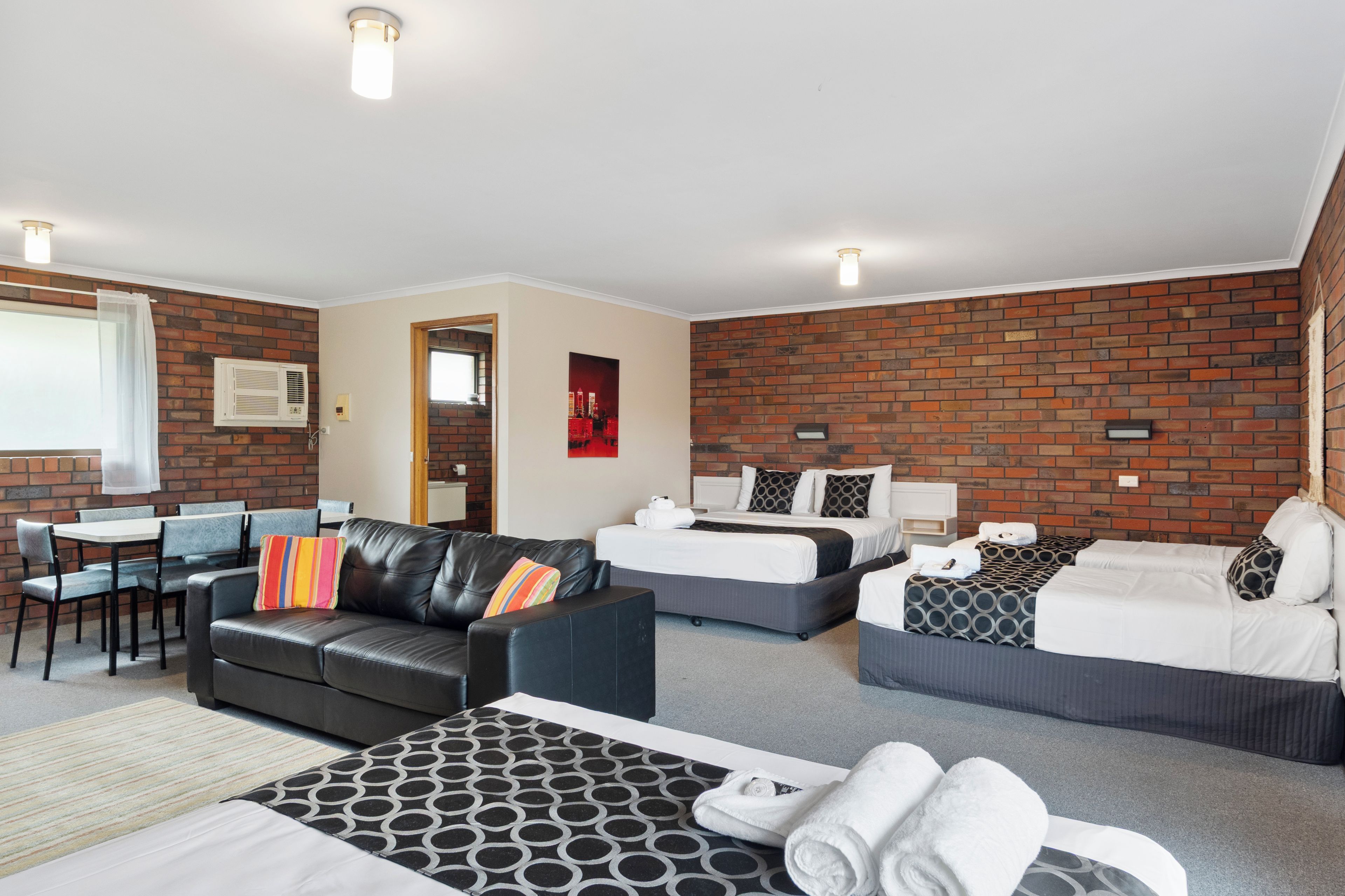 A hotel room with a brick wall, two beds, a sofa, a dining area, and a bathroom.