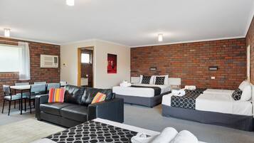 A hotel room with a brick wall, two beds, a sofa, a dining area, and a bathroom.