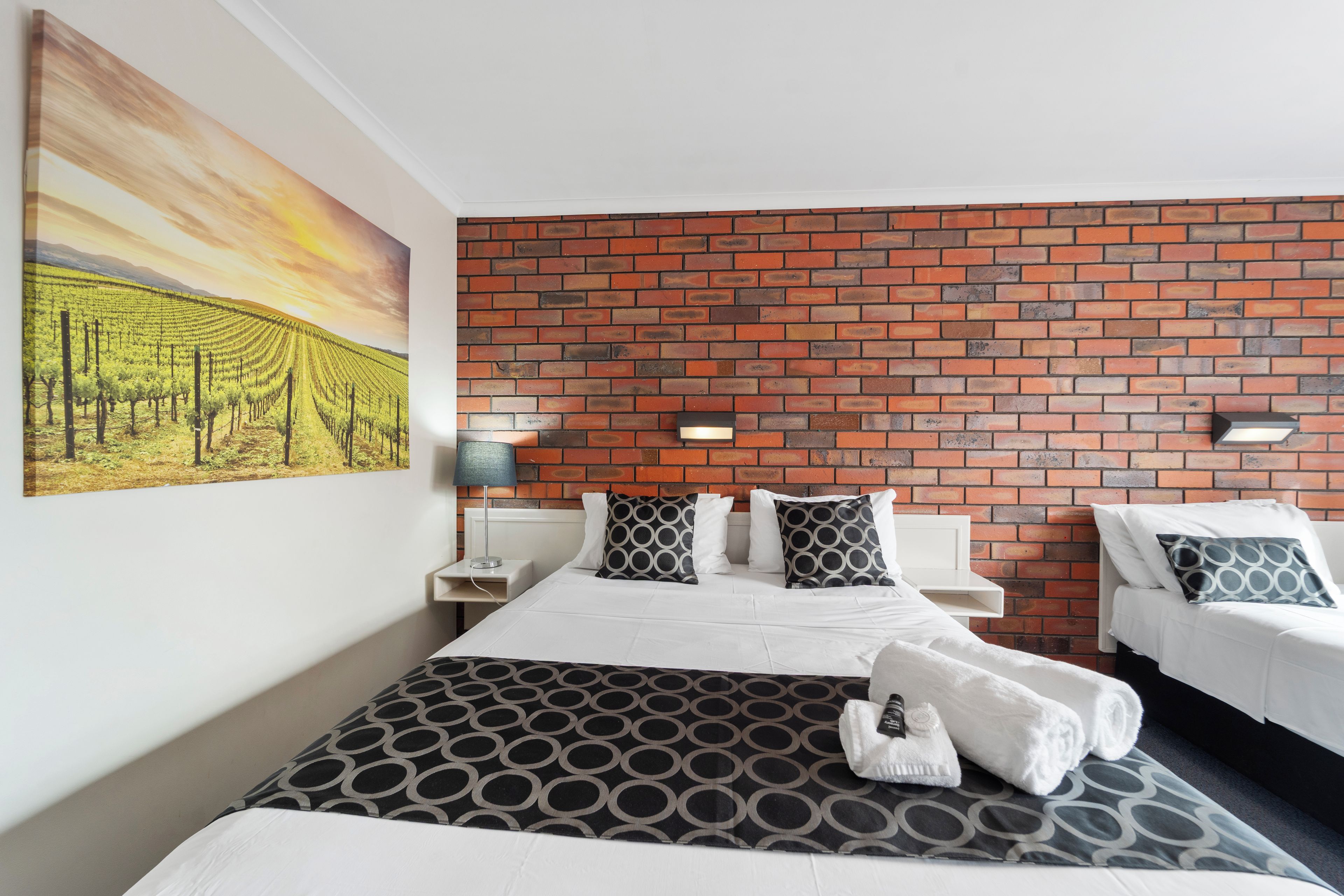 Una habitación de hotel con una pared de ladrillo, una cama con colcha estampada, dos mesitas de noche con lámparas, y un cuadro de un viñedo.