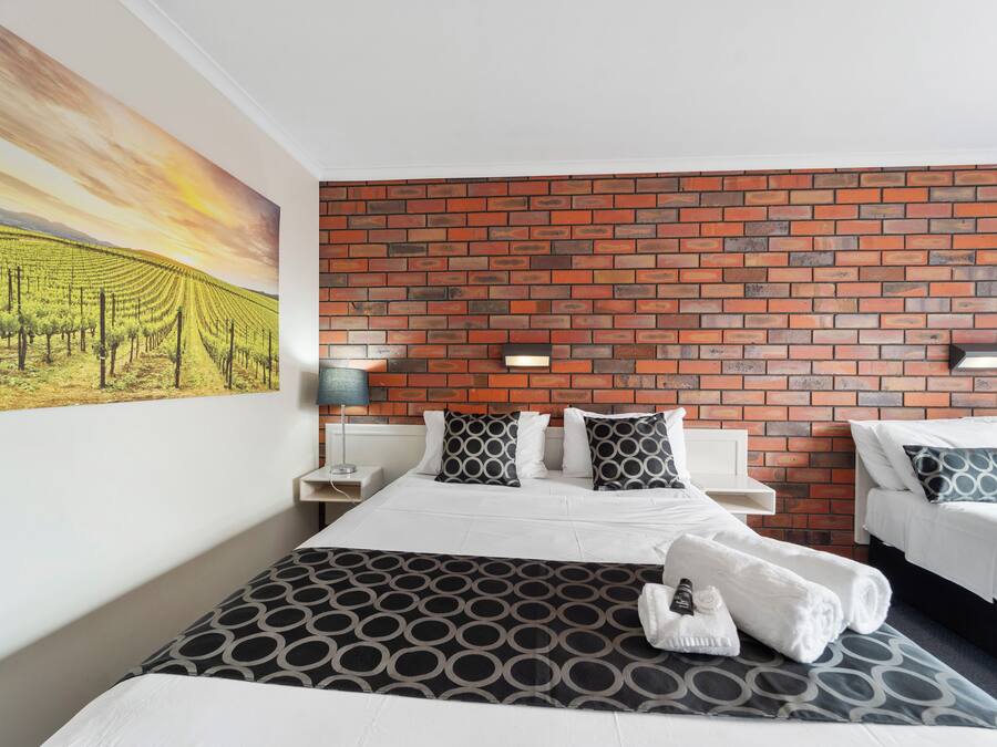 Una habitación de hotel con una pared de ladrillo, una cama con colcha estampada, dos mesitas de noche con lámparas, y un cuadro de un viñedo.