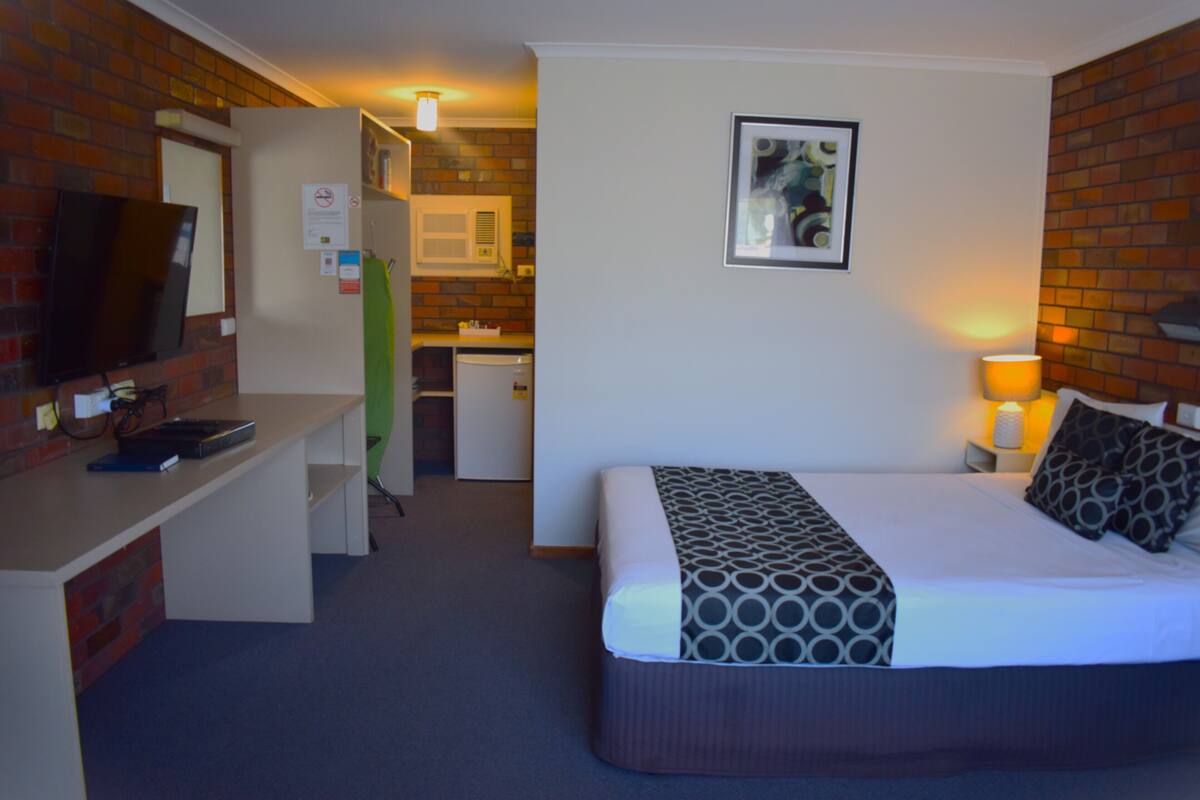 A hotel room with a bed, a desk, a television, a refrigerator, and a bathroom visible in the background.
