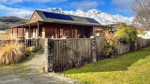 A wooden cabin with a sloped roof and solar panels, surrounded by a stone fence and a stone wall, with a mountain range in the background. - Glenorchy Lake House (Glenorchy)