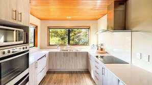 A modern kitchen with wooden cabinets, a built-in oven, a microwave, and a large window providing a view of greenery outside. - Glenorchy Lake House (Glenorchy)