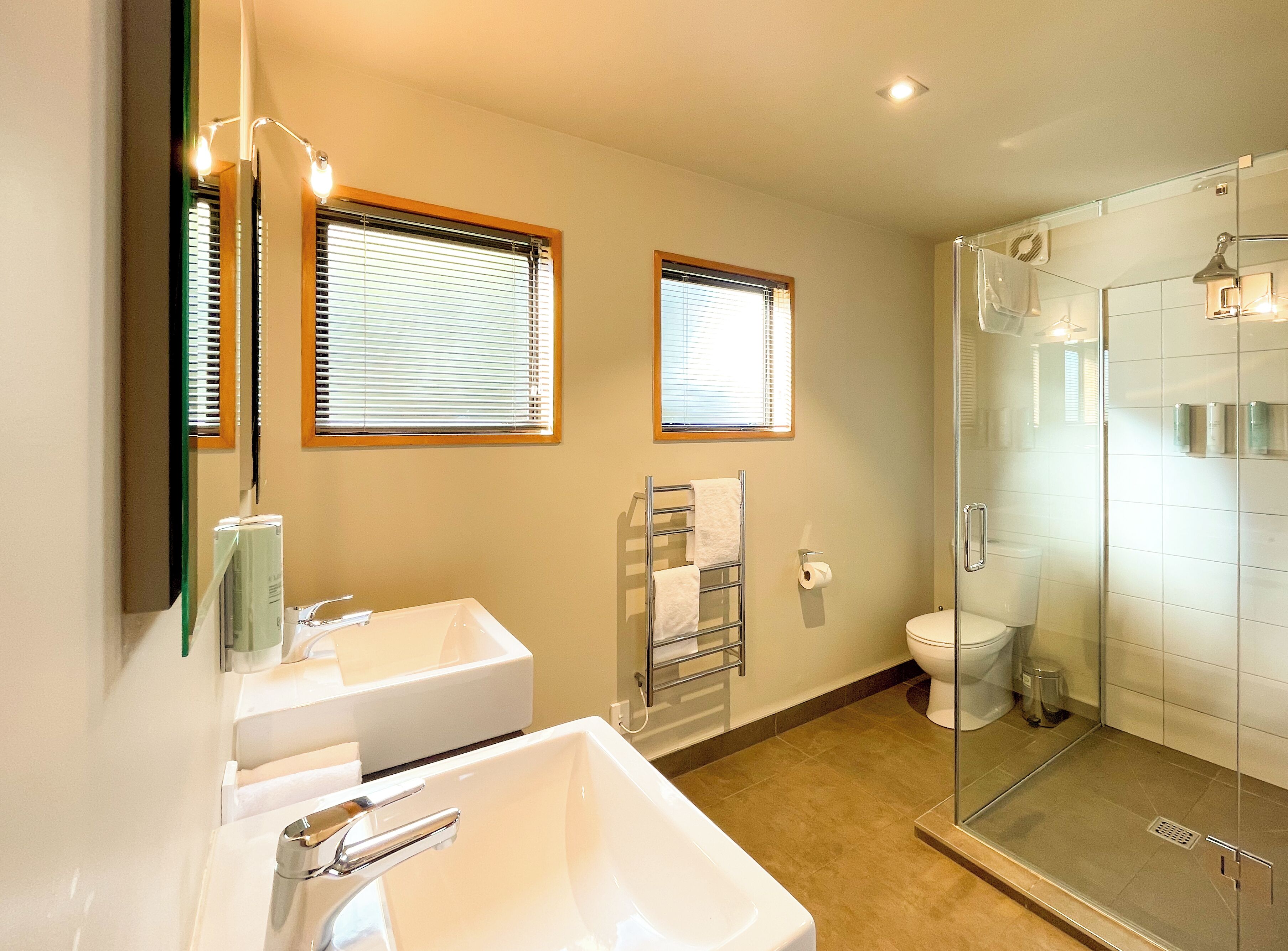 A bathroom with a glass-enclosed shower, a white sink, and two windows with blinds.