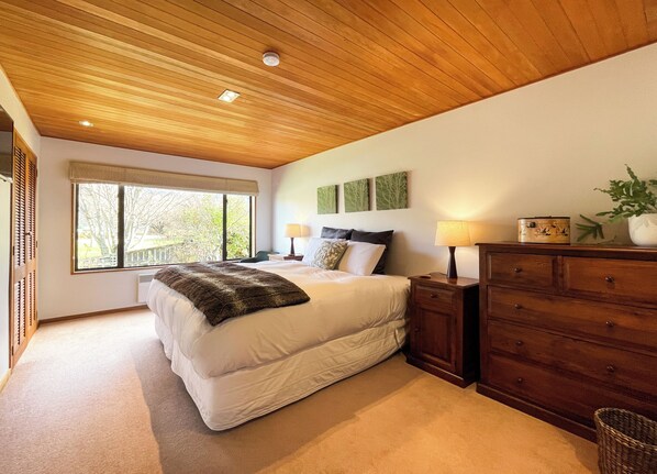 A bedroom with a large bed, wooden headboard, bedside lamps, a dresser, and a window with a view of trees.
