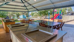 An outdoor patio with a grill station, seating area, and playground in the background. - Big4 Shepparton Park Lane (Kialla)