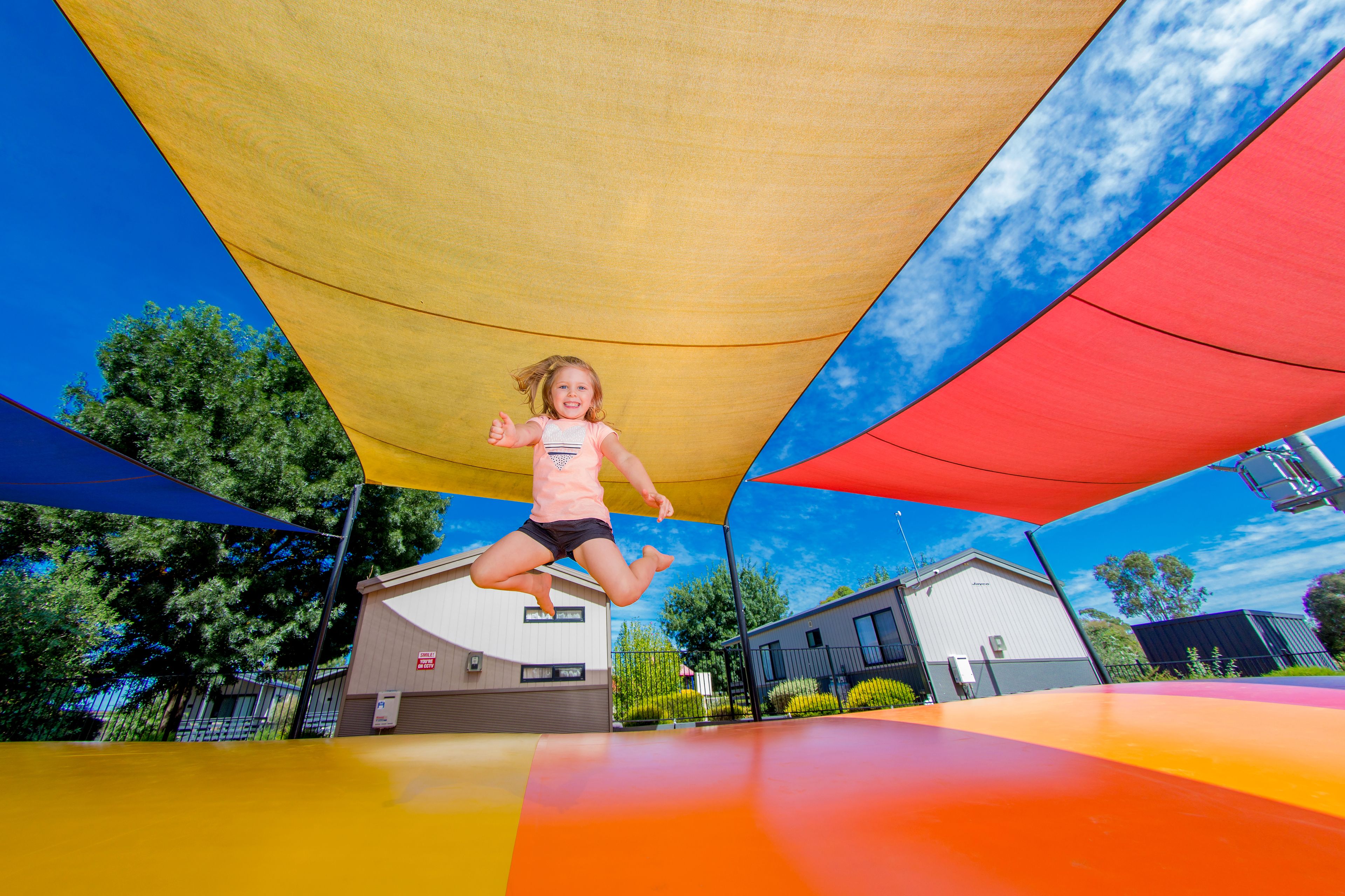 A child jumping on a trampoline under a shaded area with buildings in the background.