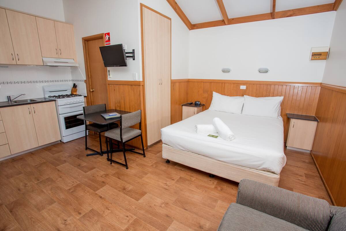 A compact room with a bed, a desk, a chair, a small table, a television, and a kitchenette.