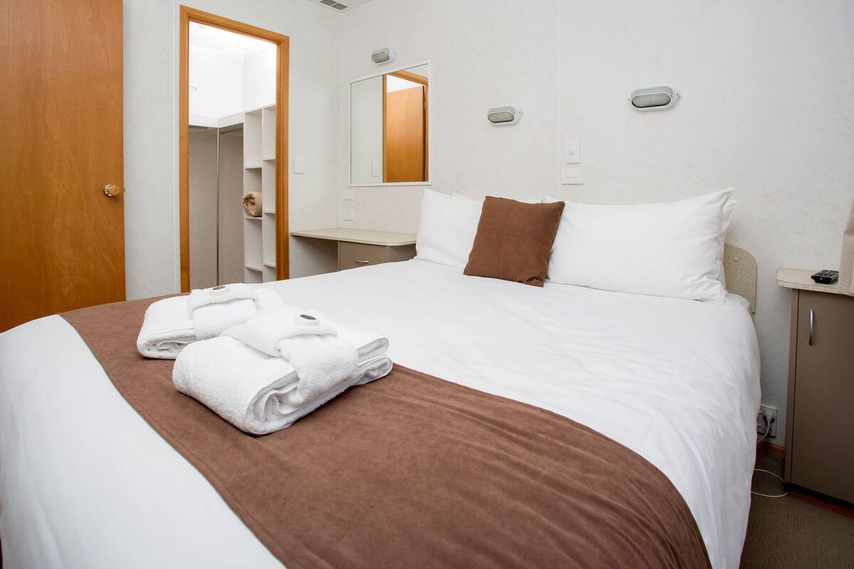 A neatly made bed with a brown accent, a wooden door, a mirror, and a bedside table with a lamp.