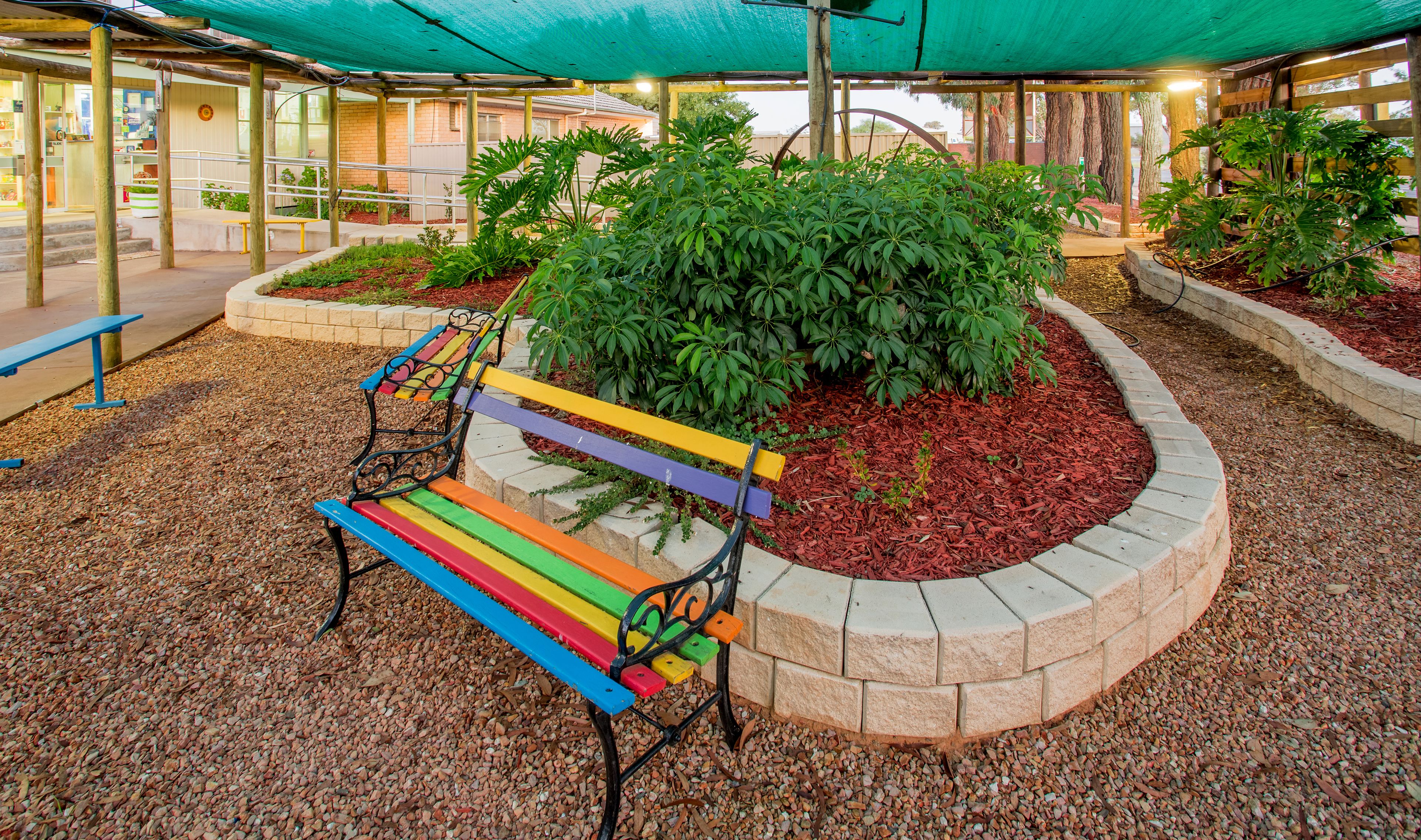 A colorful bench with a rainbow pattern, surrounded by a circular brick planter with green foliage.