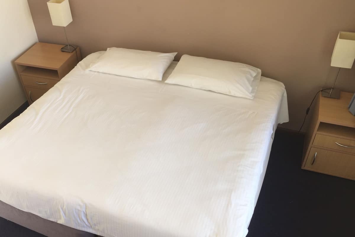 A neatly made bed with two pillows, a wooden nightstand with a lamp, and a drawer.