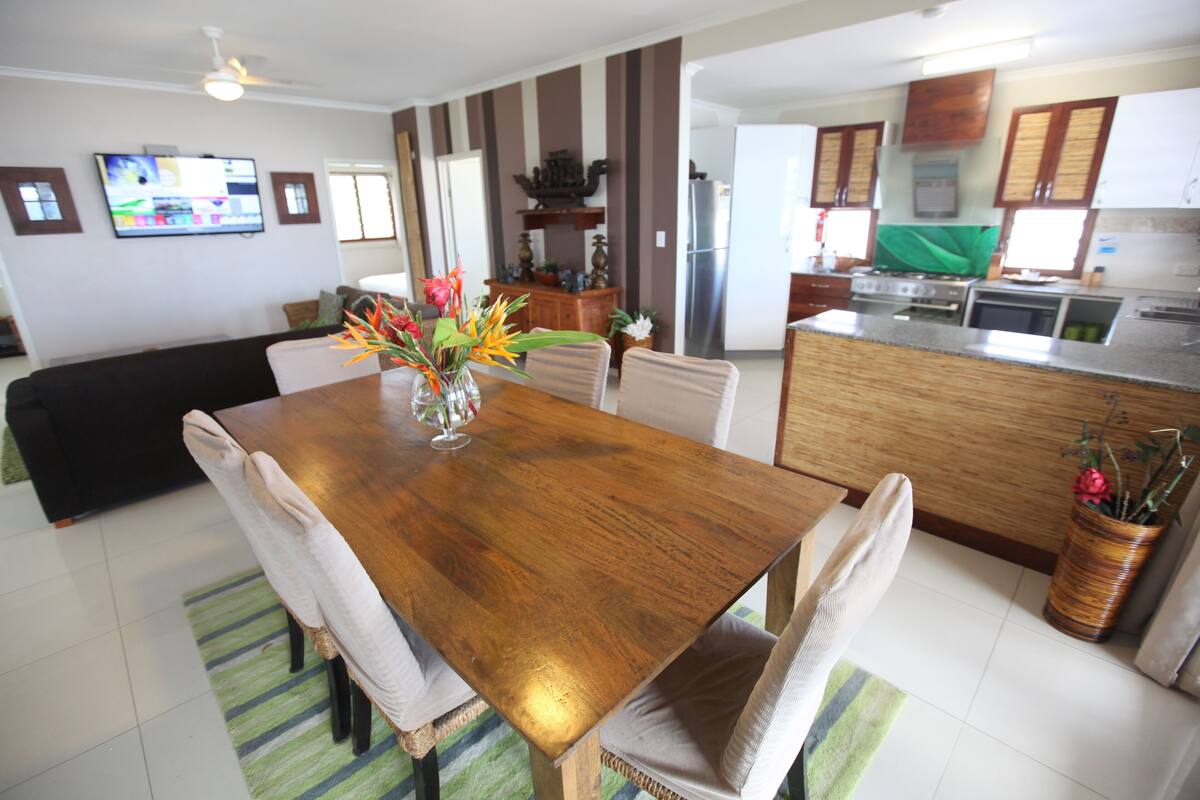 A dining area with a wooden table, chairs, and a vase of flowers. There is a kitchen in the background with appliances and cabinets.