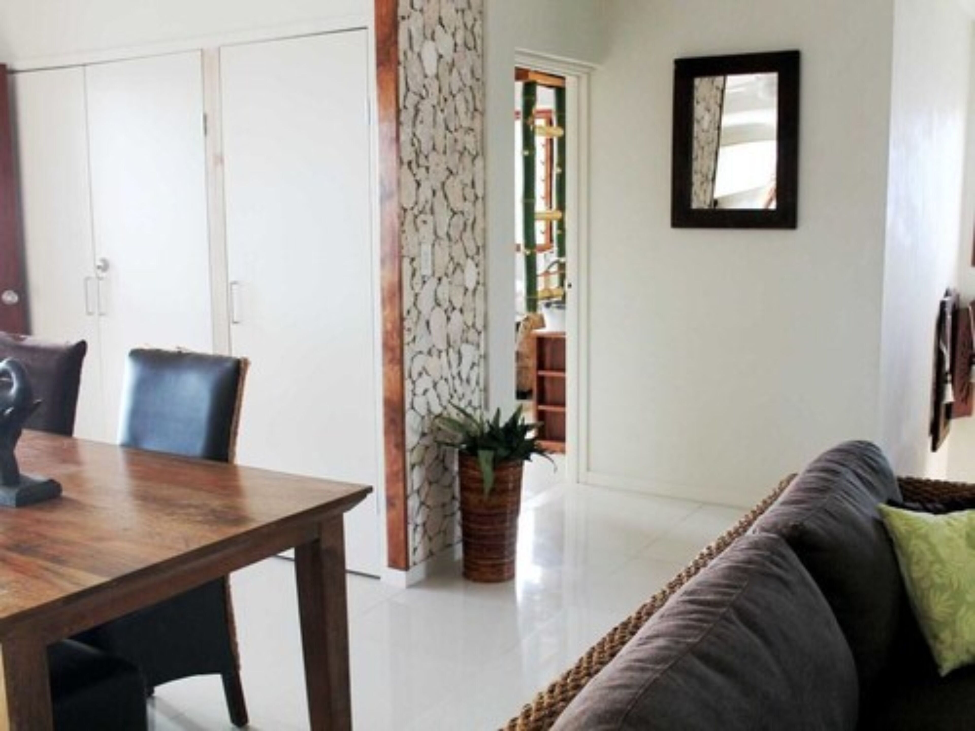 Un comedor con una mesa y sillas de madera, un sofá con cojines, una planta en maceta y un espejo en la pared.