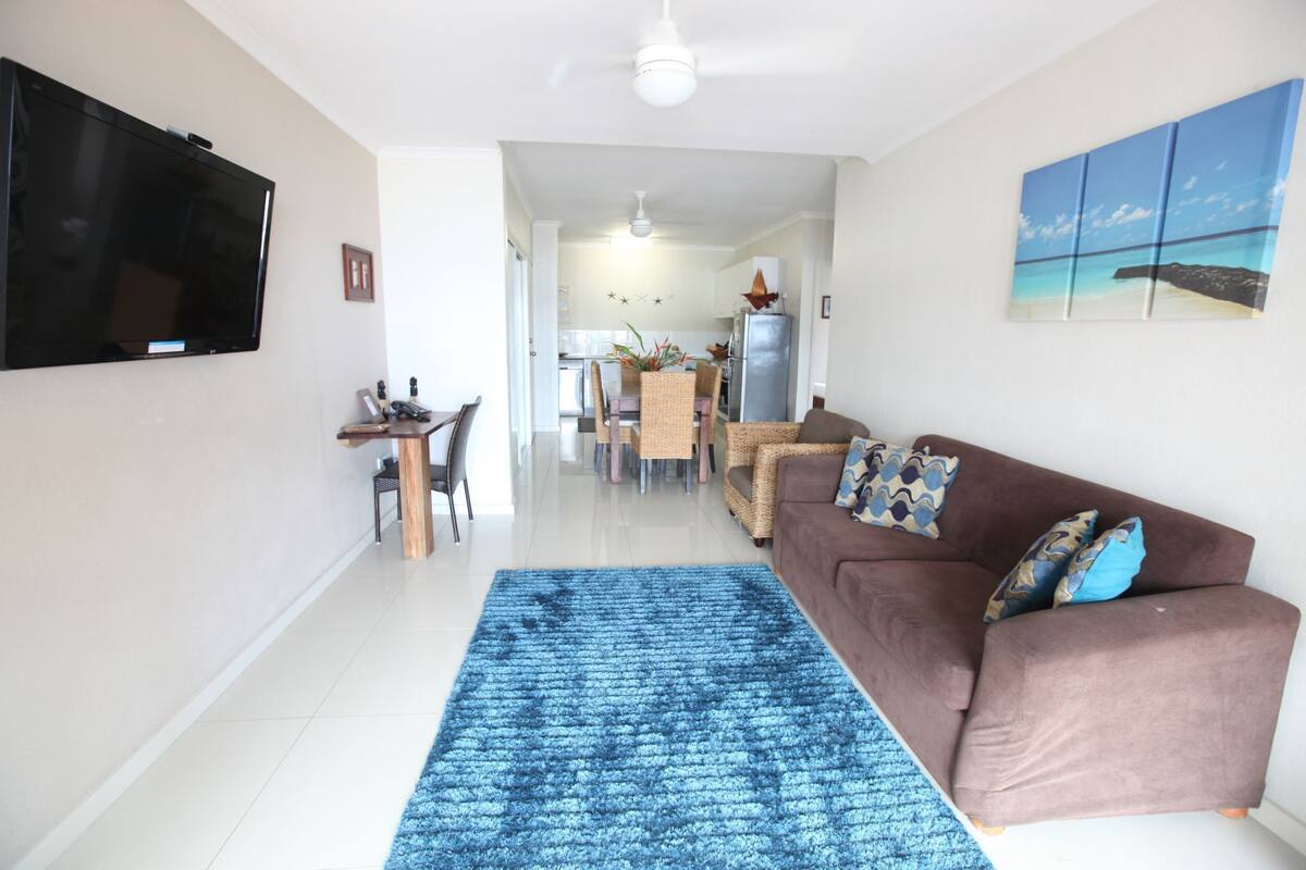 A living room with a brown sofa, a blue rug, a flat-screen TV, and a dining area with a table and chairs.