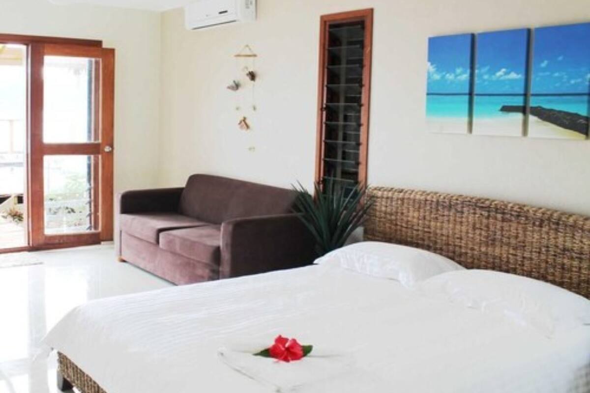 A bedroom with a bed, a brown sofa, a wicker chair, a window with a view, and a wall-mounted air conditioner.