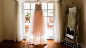 A wedding dress hanging in front of a window with curtains, a mirror, and a potted plant. - Lakeside Cottage (Kilsyth South)