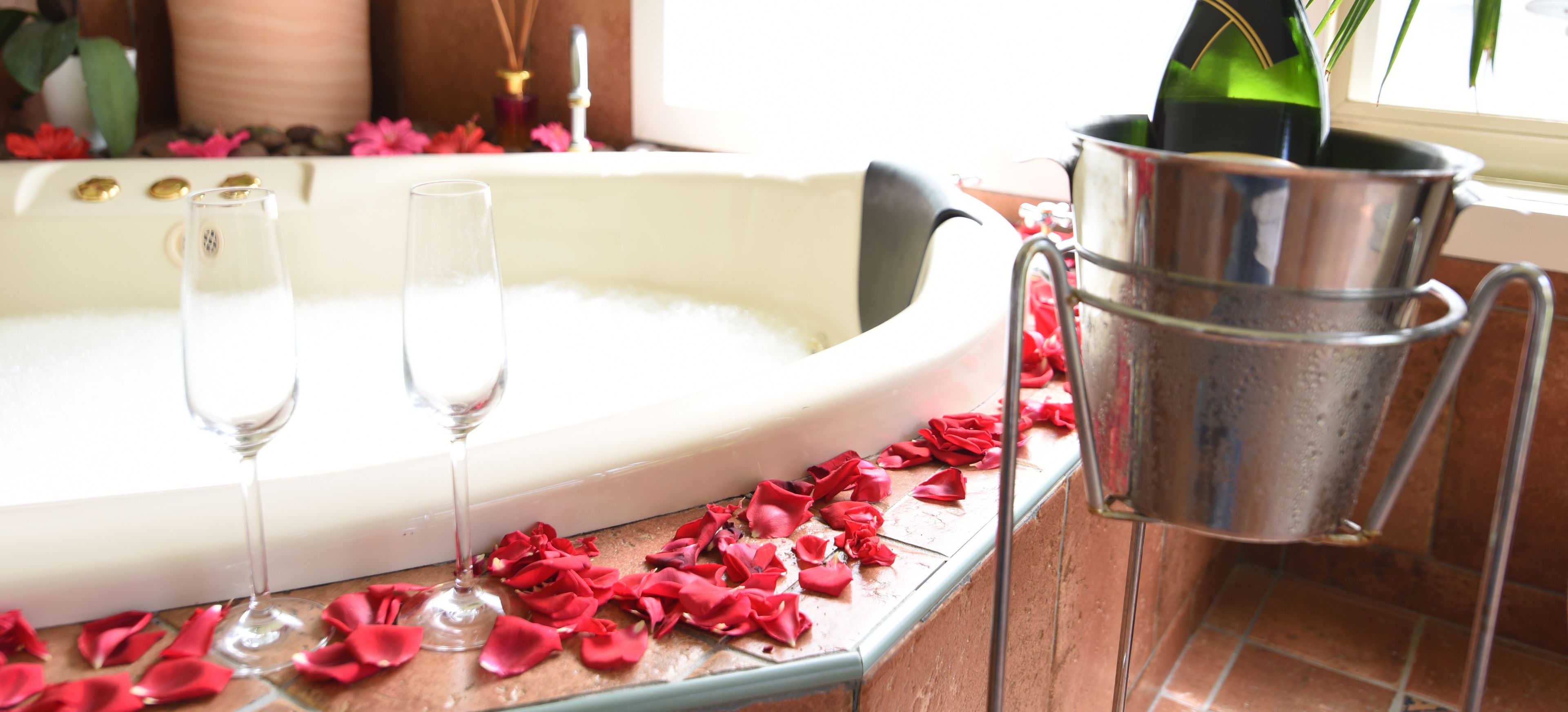 A bathtub filled with water, two empty glasses, and a champagne bottle in a bucket.