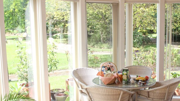 A conservatory with wicker chairs and a glass table, surrounded by potted plants and a view of a garden. - Lakeside Cottage (Kilsyth South)