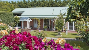 A house with a green roof, surrounded by a garden with various flowers and a small tree. - Lakeside Cottage (Kilsyth South)