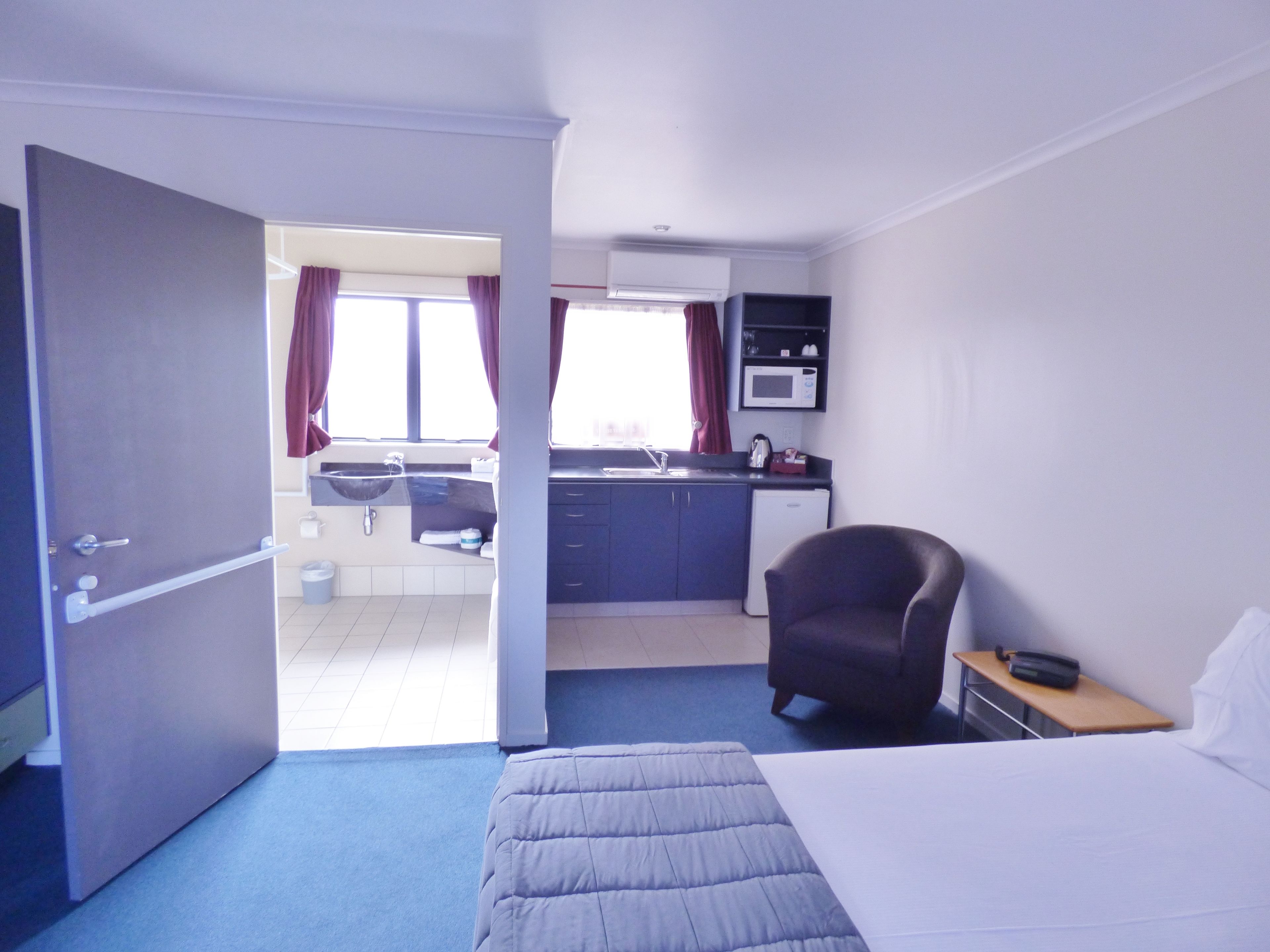 Habitación de hotel con cama, escritorio con silla, kitchenette y baño visible a través de una puerta abierta.