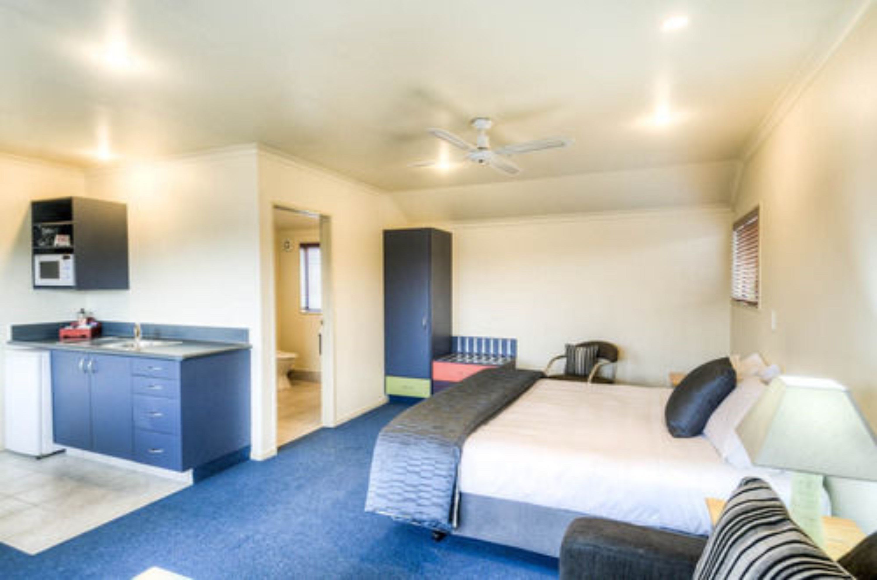 A compact hotel room with a kitchenette, a bed with a headboard, a bedside table with a lamp, and a ceiling fan.