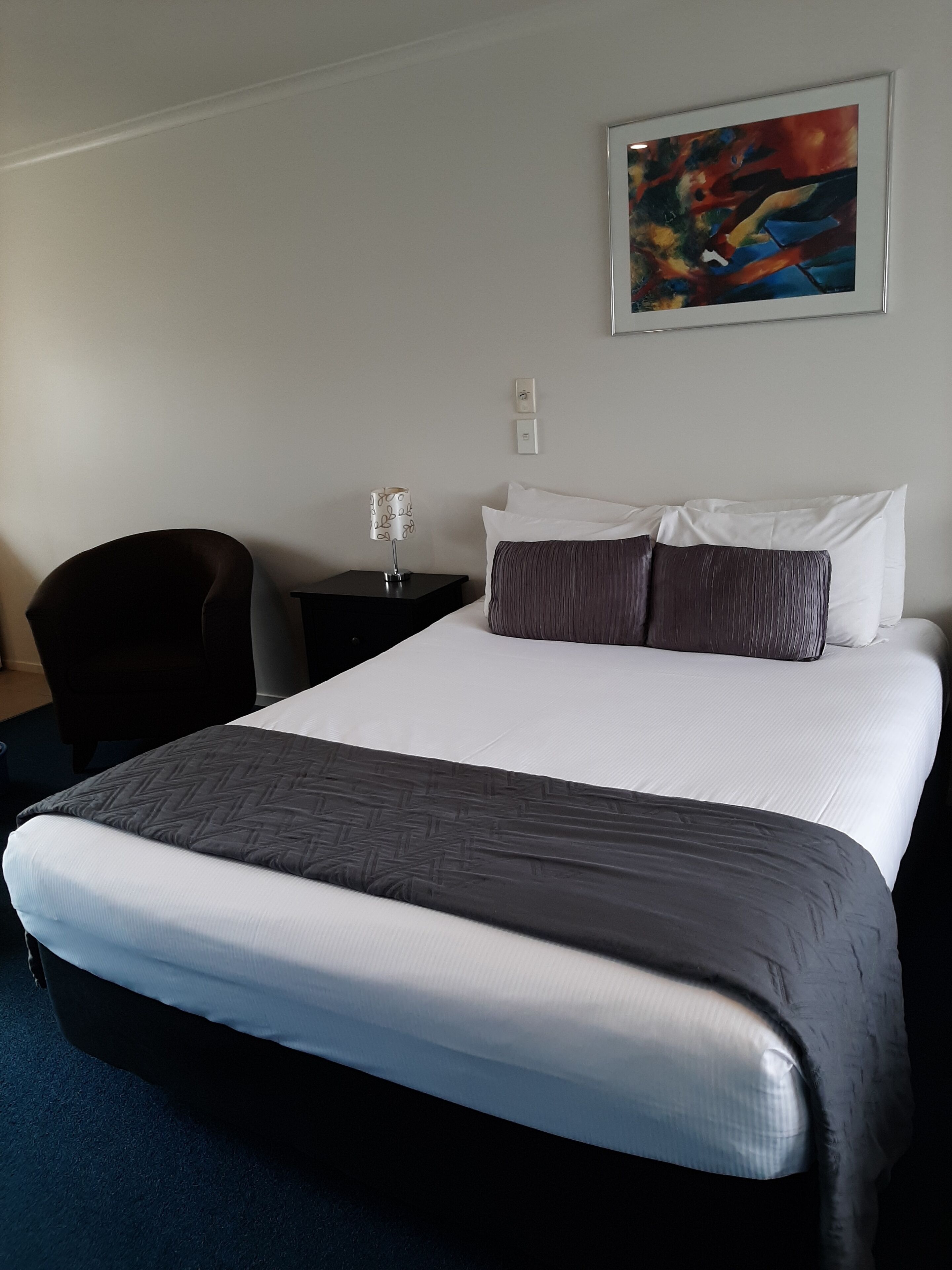 A hotel room with a large bed, a nightstand, a lamp, a chair, and a painting on the wall.