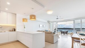 Et moderne køkken med hvide skabe, en central ø og et spiseområde med bord og stole. Et stort vindue giver udsigt til havet.