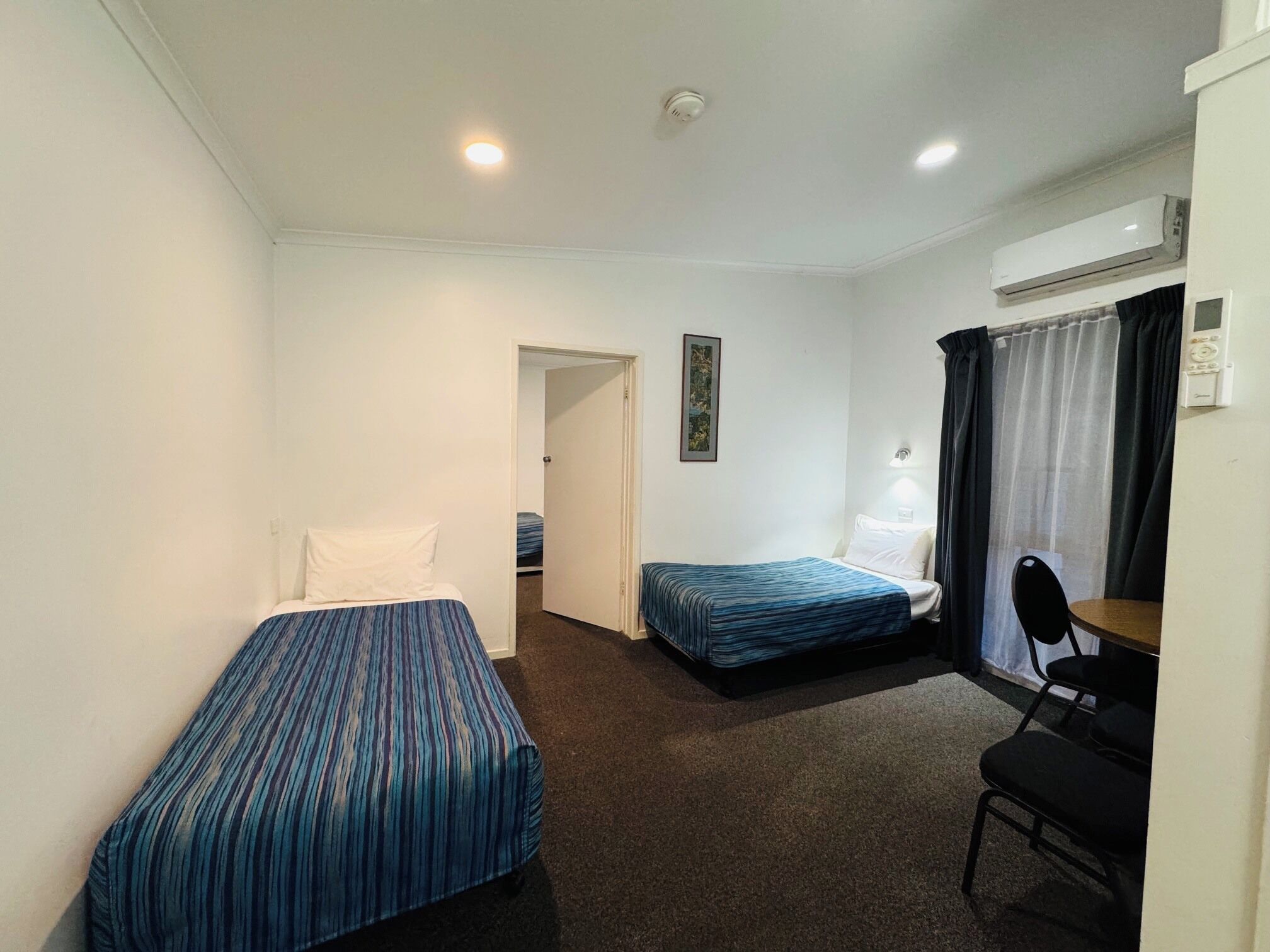 Ein Zimmer mit zwei Einzelbetten, einem Schreibtisch mit Stuhl, einem kleinen Tisch, einem Fenster mit Vorhängen und einer Klimaanlage.