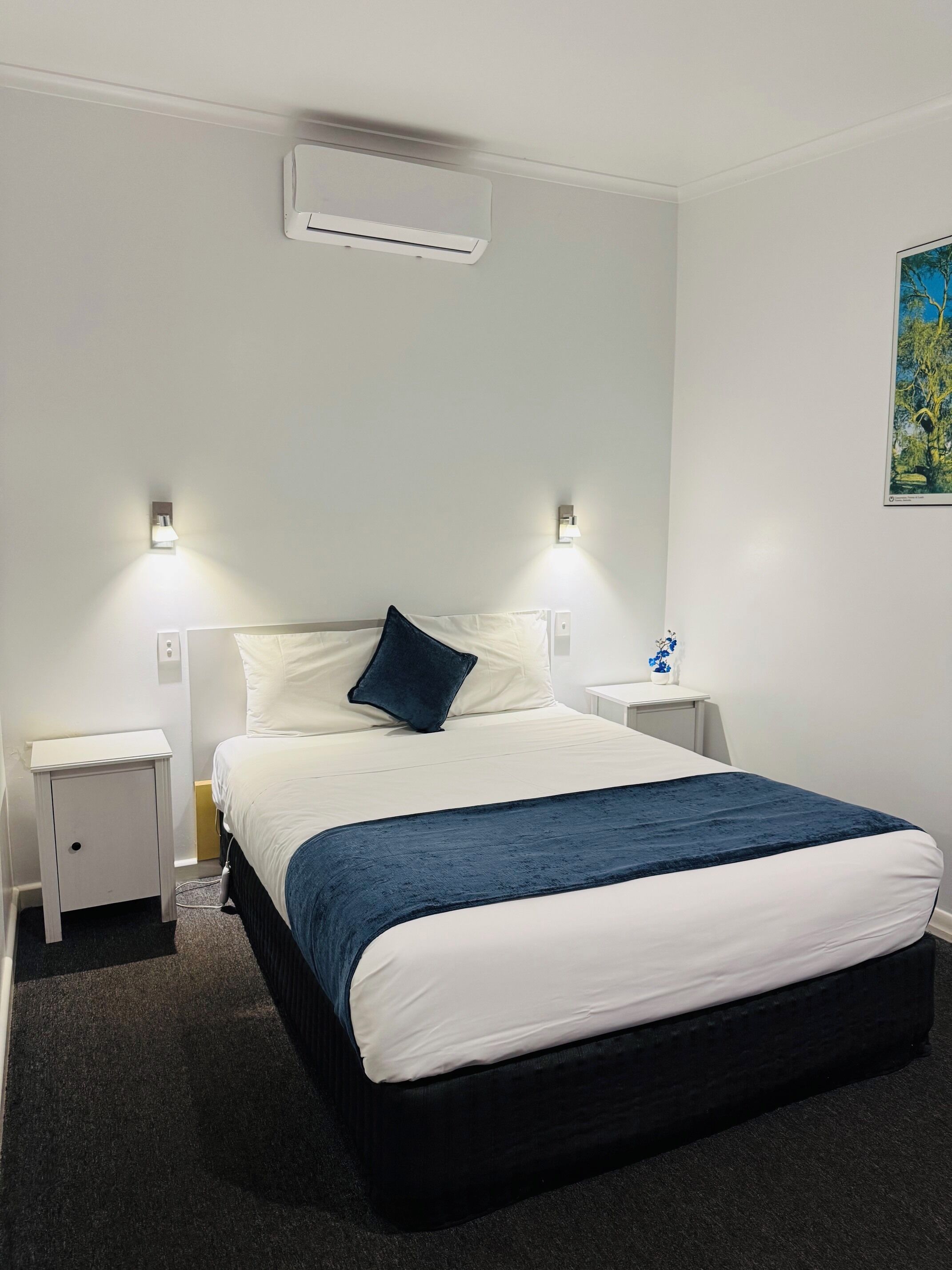 A hotel room with a bed, two bedside tables, a nightstand, an air conditioning unit, and a wall-mounted light fixture.
