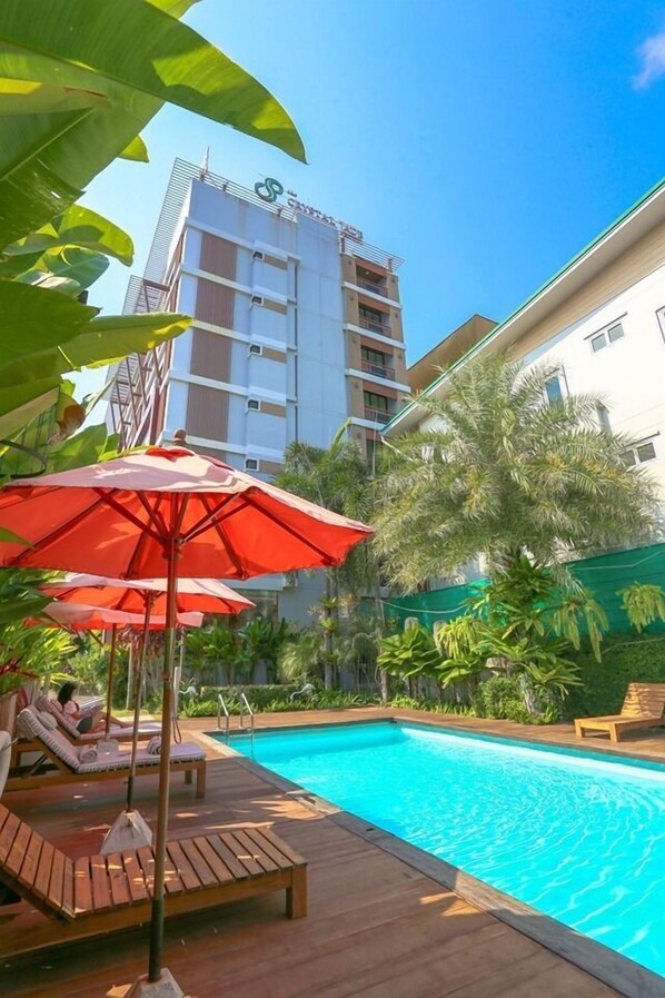 A swimming pool with wooden loungers and red umbrellas, surrounded by greenery and a tall building in the background. - Crystal Jade Hotel (Rayong)