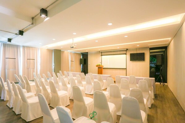 Un salón de conferencias con filas de sillas blancas, un atril, una pantalla de proyector y un amplio ventanal con cortinas.