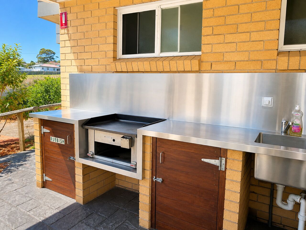 An outdoor kitchen with a stainless steel countertop, a built-in grill, and a wooden cabinet labeled 'RUBBISH'.