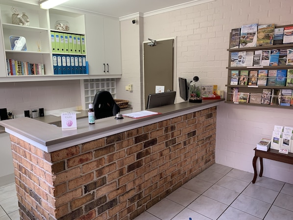 A reception desk with a brick base, a computer, and a telephone. There are shelves with books and binders. A basket and a small plant are on the desk. - Ulladulla Harbour Motel (Ulladulla)
