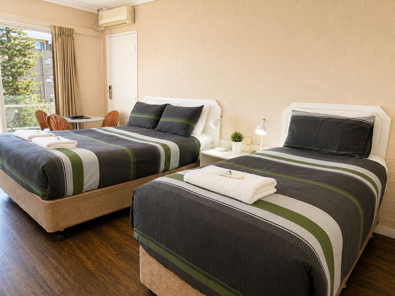 Quarto de hotel com duas camas, uma mesinha, uma cadeira e uma janela com cortinas.