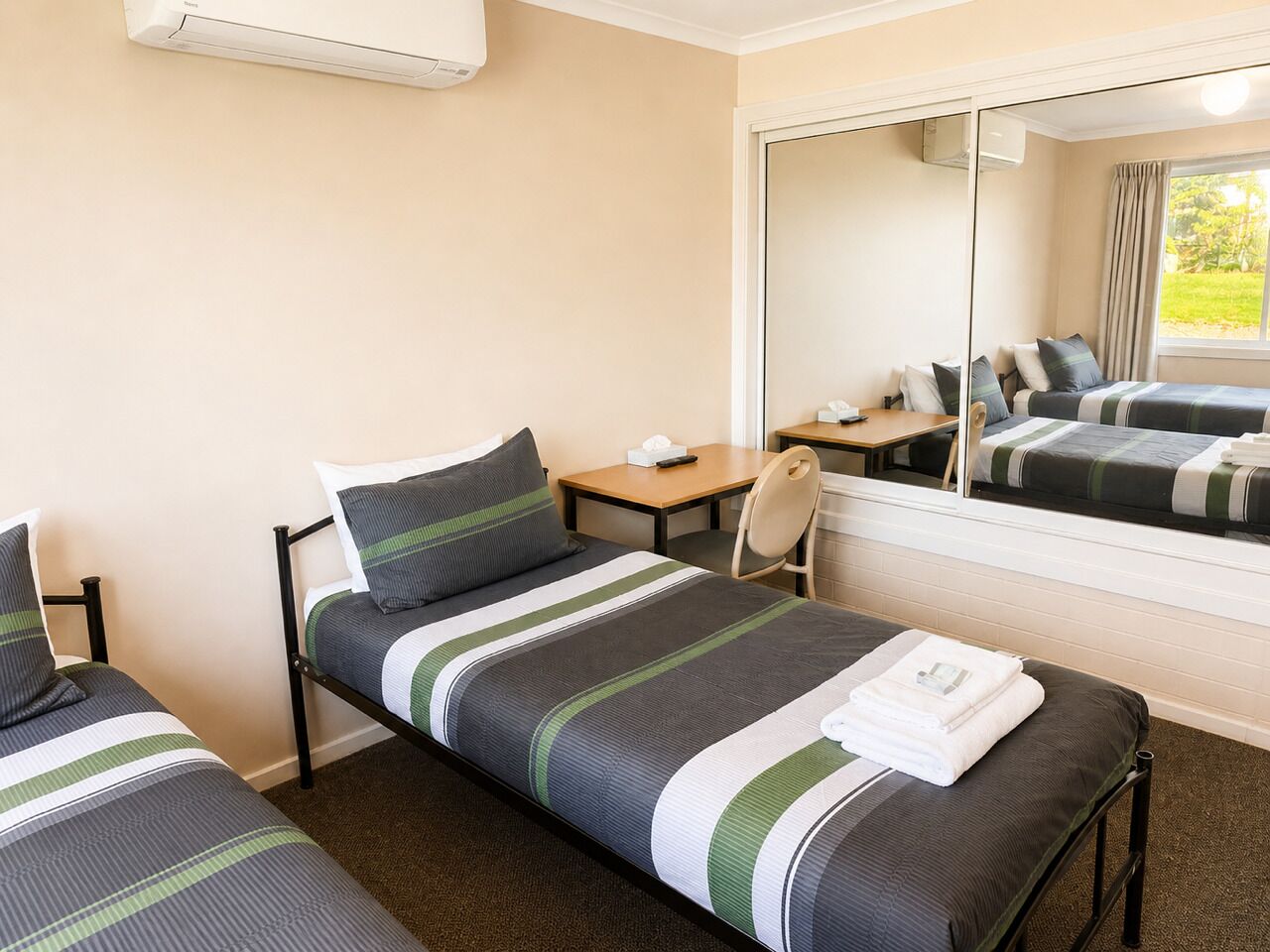 Habitación doble con dos camas individuales, una mesita, una silla, un espejo y una ventana con cortinas.