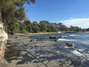 A rocky shoreline with clear blue water, trees, and a distant building. - Ulladulla Harbour Motel (Ulladulla)