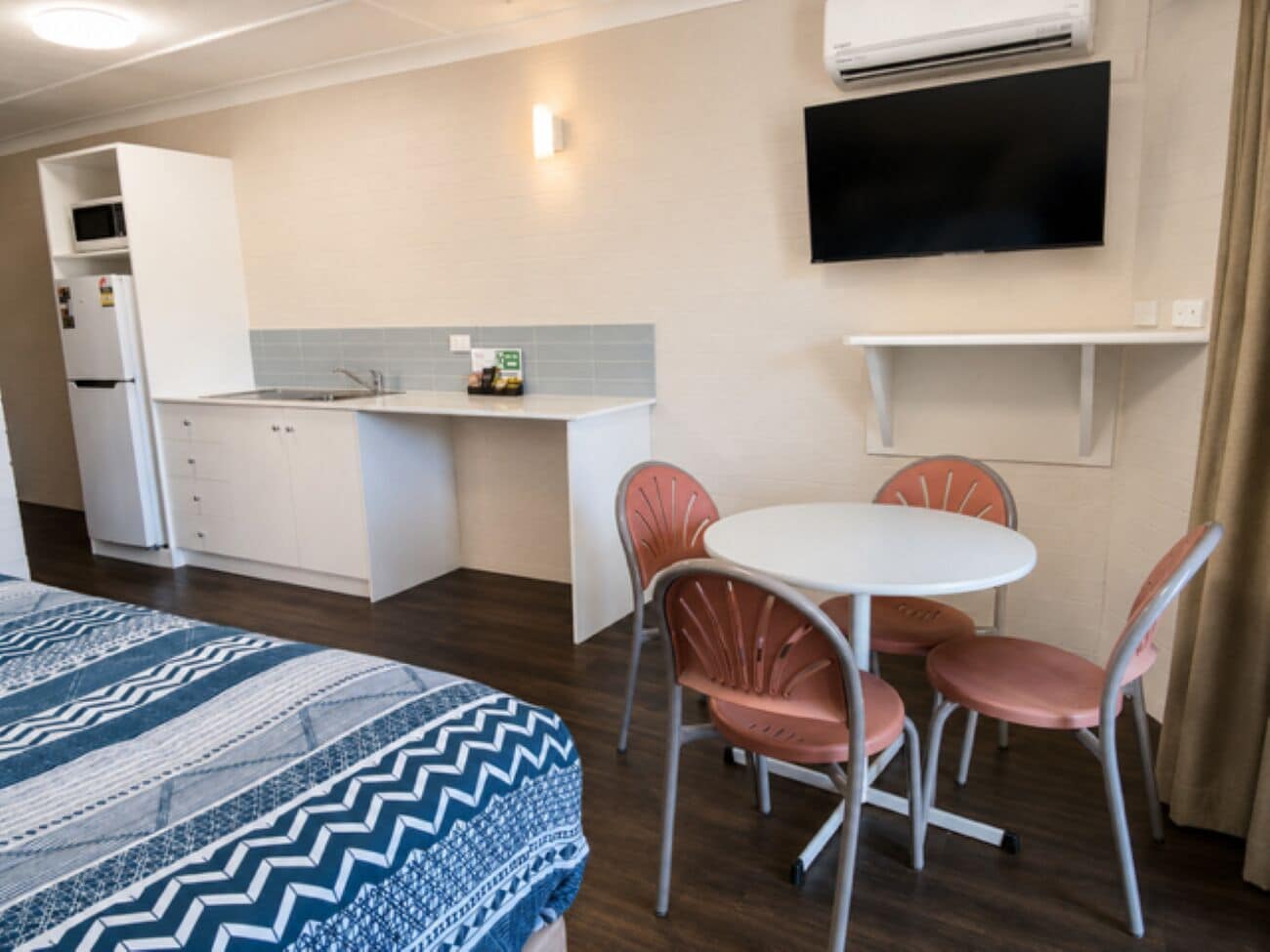 Quarto de hotel compacto com cozinha compacta, cama com roupa de cama estampada, área de jantar com mesa e cadeiras, e televisão de tela plana fixada na parede.