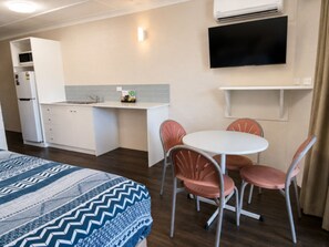 Habitación de hotel compacta con kitchenette, cama con ropa de cama estampada, zona de comedor con mesa y sillas, y televisor de pantalla plana montado en la pared.