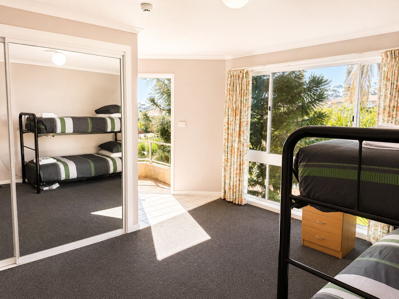 A room with a bunk bed, a single bed, a wooden nightstand, and a large window with curtains.