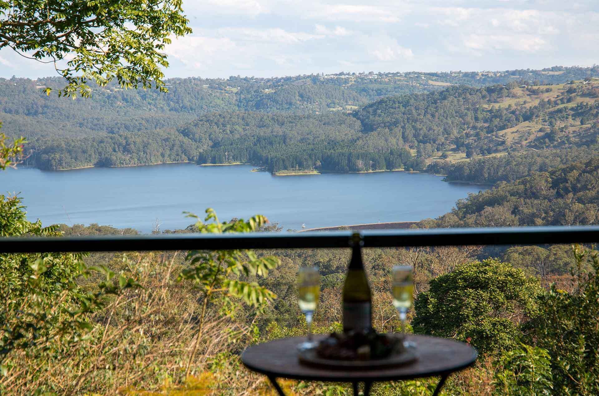 A table with a bottle and glasses, overlooking a lake surrounded by forested hills.