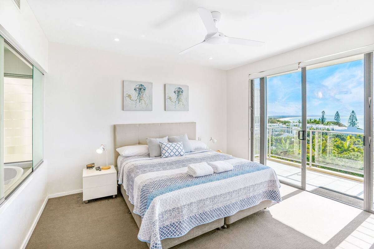 A modern bedroom with a large bed, a nightstand, a ceiling fan, and a balcony with a view of the ocean and greenery.