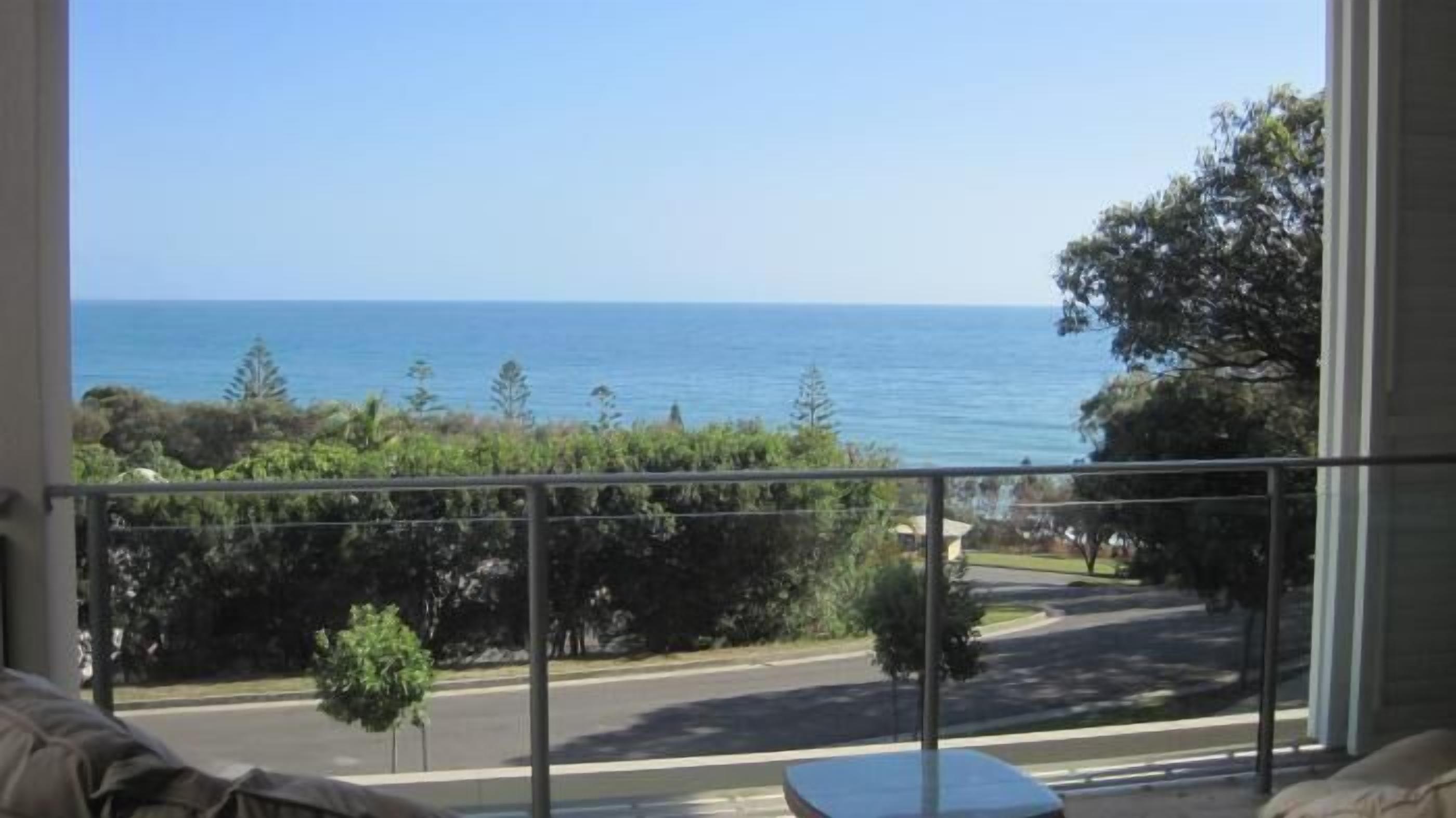 A balcony with a view of the ocean and a road with trees and a small building in the distance.