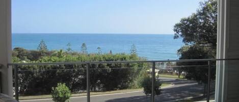 A balcony with a view of the ocean and a road with trees and a small building in the distance.