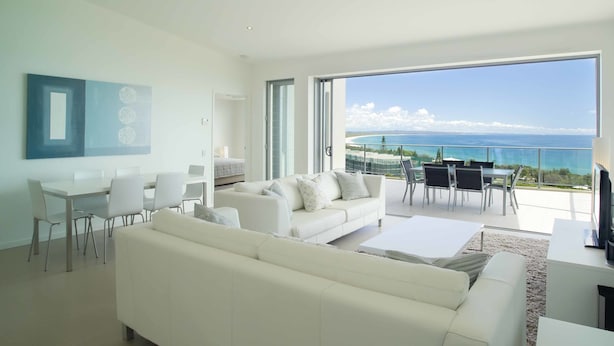 A spacious living room with a white sofa, dining table, and chairs. A large window offers a view of the ocean.
