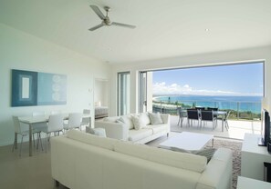 Un salon spacieux avec un canapé blanc, une table à manger et des chaises. Une grande fenêtre offre une vue sur l’océan.