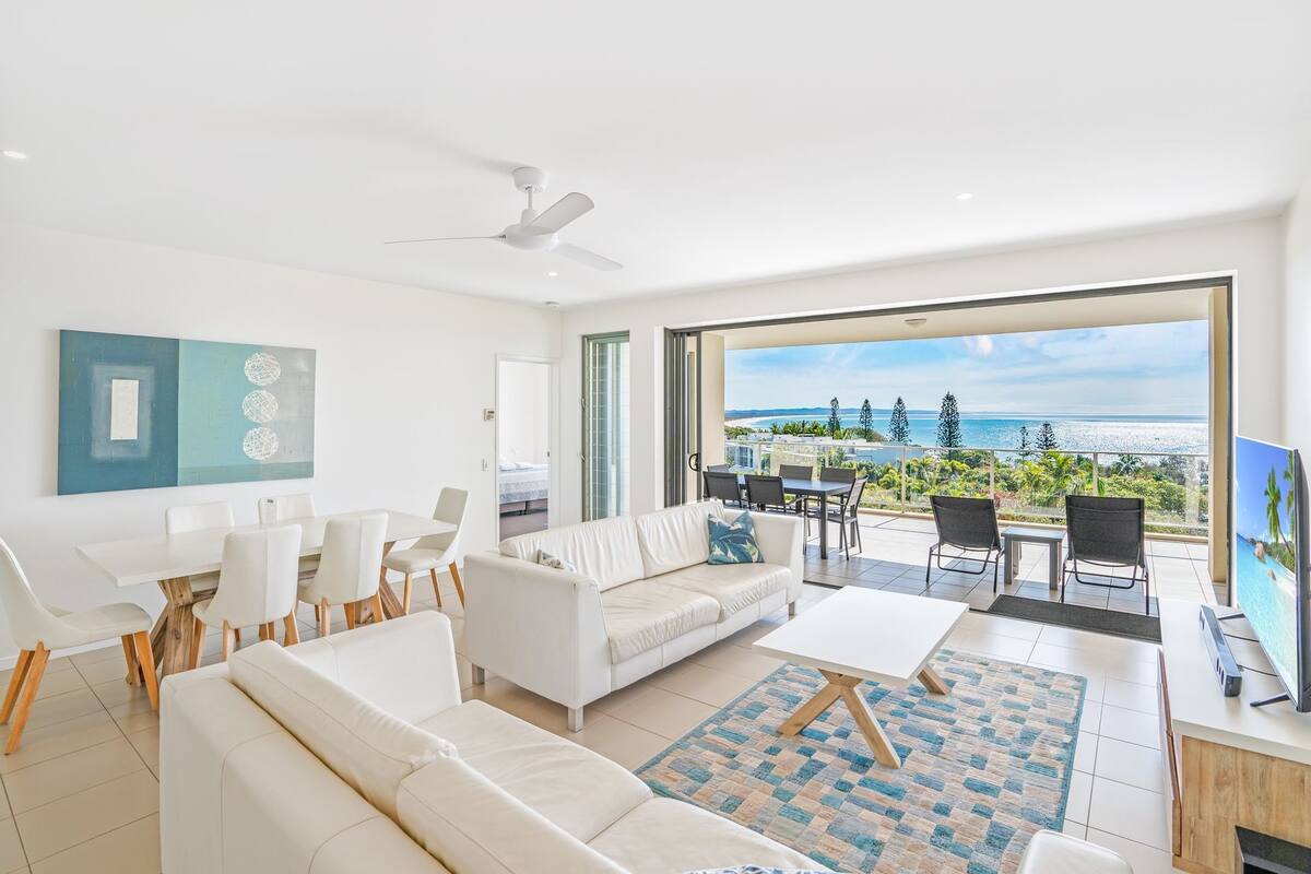 A modern living room with a white sofa, a dining table, and chairs. There is a large window with a view of the ocean and palm trees.